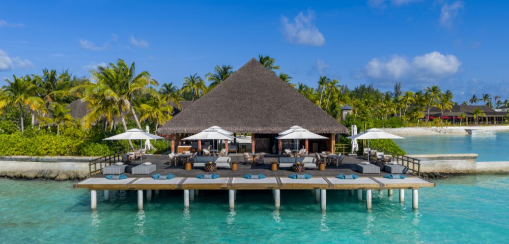 A tropical resort with thatched-roof building on stilts over turquoise water. Surrounding the building are palm trees and clear skies, evoking relaxation.