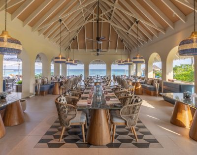 Open-air restaurant with high ceilings and ocean view. Features wicker chairs, wooden tables, and pendant lights, creating a relaxed and inviting atmosphere.