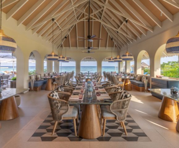 Open-air restaurant with high ceilings and ocean view. Features wicker chairs, wooden tables, and pendant lights, creating a relaxed and inviting atmosphere.