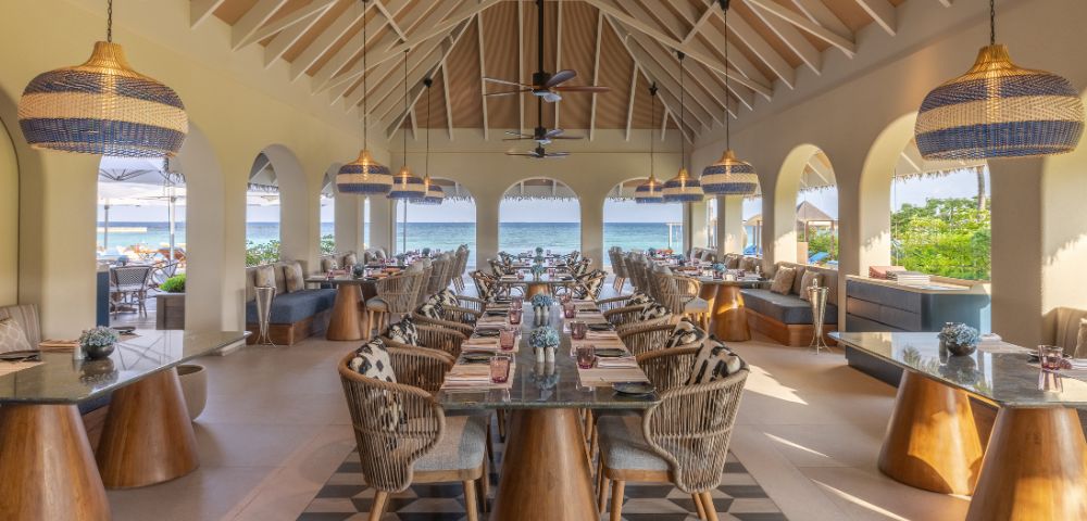 Open-air restaurant with high ceilings and ocean view. Features wicker chairs, wooden tables, and pendant lights, creating a relaxed and inviting atmosphere.