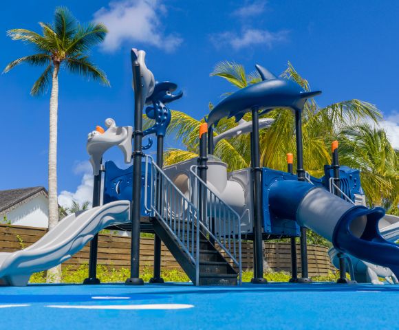 Children's playground with blue slides and climbing structures under a clear sky, surrounded by palm trees, conveying a fun and tropical vibe.