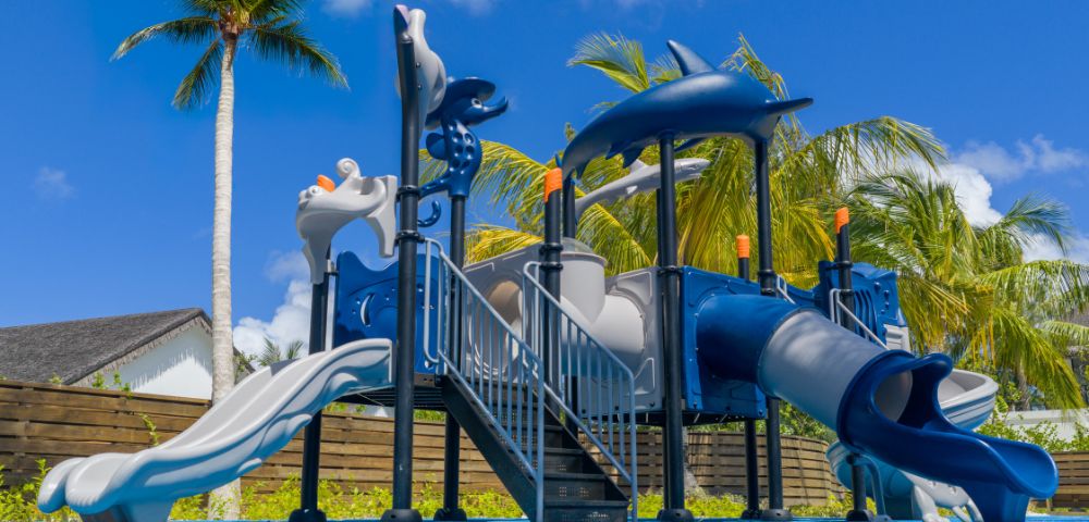 Children's playground with blue slides and climbing structures under a clear sky, surrounded by palm trees, conveying a fun and tropical vibe.