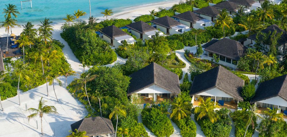 Aerial view of a tropical beach resort with white villas surrounded by lush greenery and palm trees on a pristine sandy beach beside turquoise water.