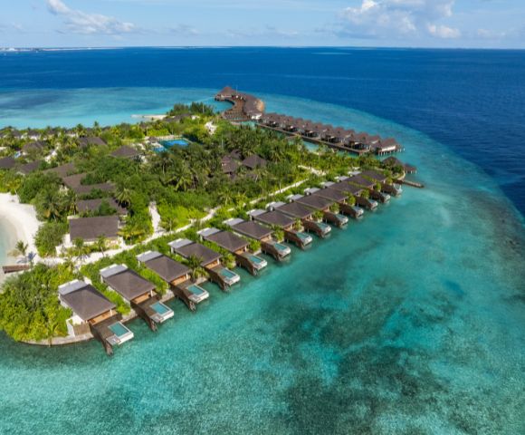 Aerial view of a tropical island resort with thatched-roof villas over turquoise water. Lush greenery, sandy beach, and a peaceful, serene atmosphere.