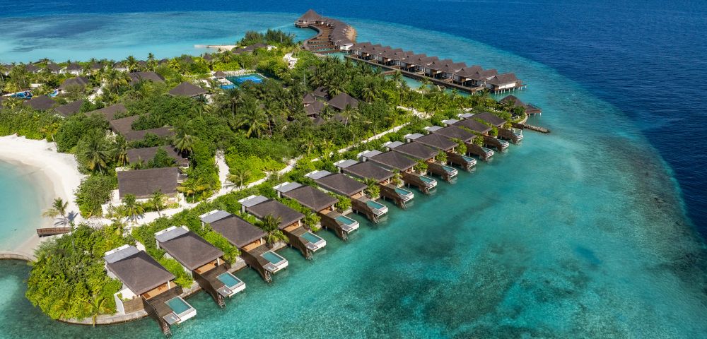 Aerial view of a tropical island resort with thatched-roof villas over turquoise water. Lush greenery, sandy beach, and a peaceful, serene atmosphere.