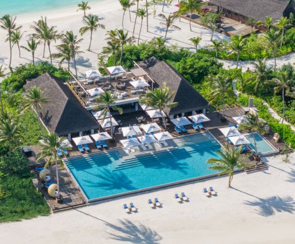 Aerial view of a luxury beachfront resort with a large pool surrounded by palm trees and deck chairs. The serene setting conveys relaxation and tropical paradise.