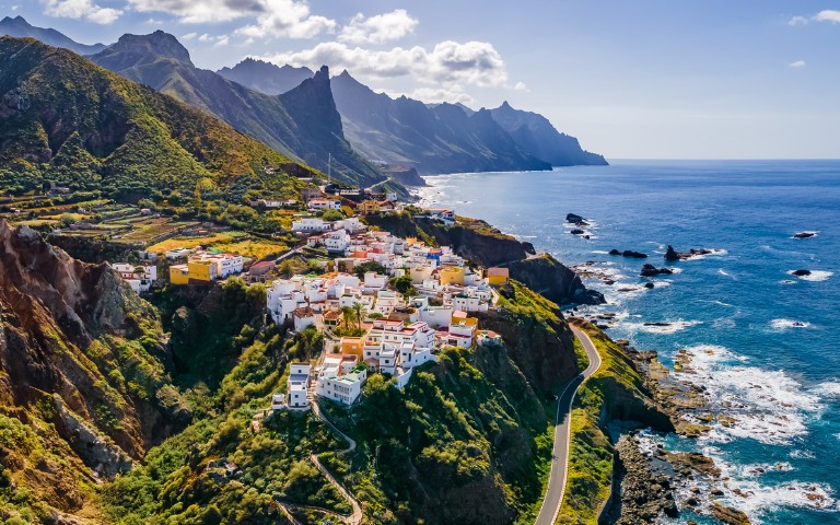 A picturesque coastal village with colorful houses atop a lush, green cliff. Mountains rise in the background, and the vast ocean stretches under a bright blue sky.