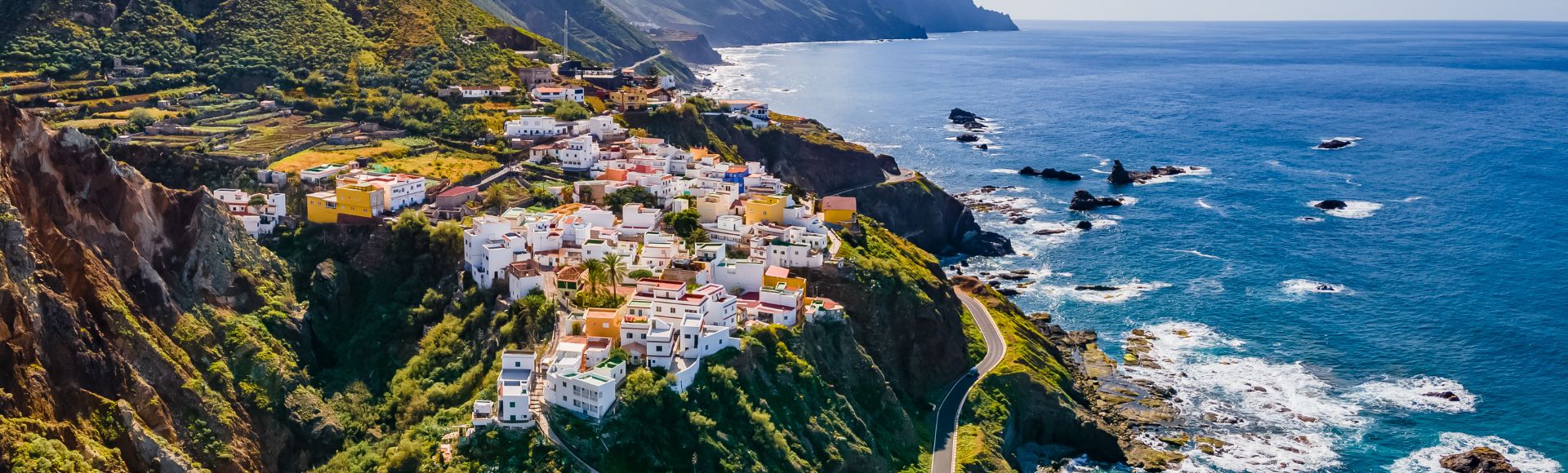 A picturesque coastal village with colorful houses atop a lush, green cliff. Mountains rise in the background, and the vast ocean stretches under a bright blue sky.