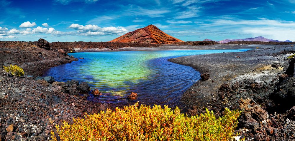 A vibrant landscape features a serene blue lagoon bordered by volcanic rocks and bright green vegetation, with a striking red-orange mountain under a vivid blue sky.