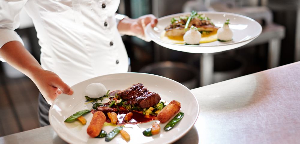A waiter in a white shirt presents two gourmet plates: one with steak, vegetables, and sauce, the other with a savory dish and garnishes.