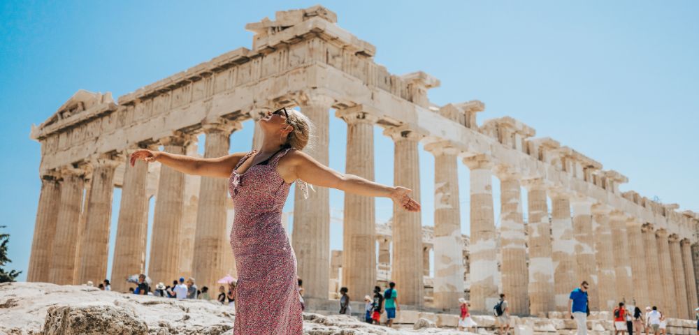 A woman stands in front of the Parthenon, arms outstretched, enjoying the sun on a clear day amidst a crowd of tourists.