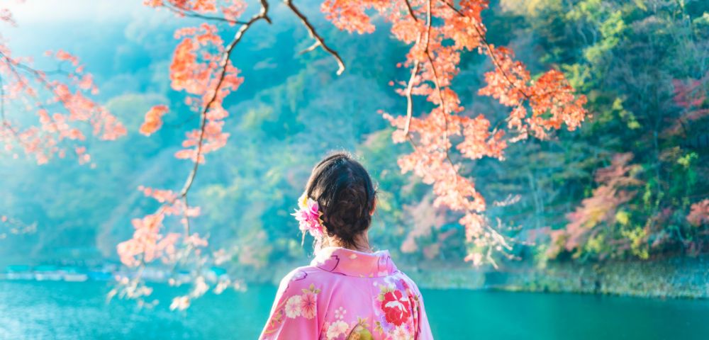 A woman in a floral kimono stands by a serene lake, surrounded by vibrant autumn leaves and lush greenery.