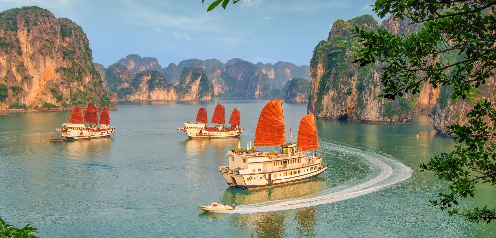 Three traditional boats with red sails navigate a serene bay, surrounded by towering limestone cliffs and lush greenery under a blue sky.