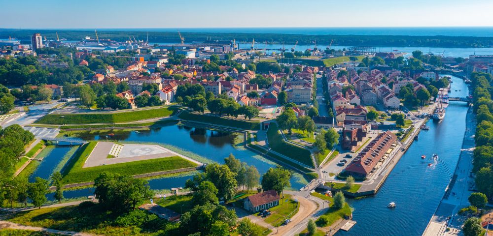 Aerial view of a vibrant coastal town with a river winding through it. Lush greenery surrounds historic buildings. The harbor and ocean are visible beyond.