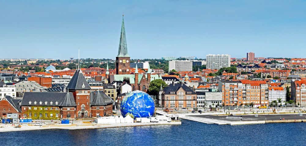 Aerial view of Aarhus, Denmark, featuring a striking blue globe art installation by the waterfront. Historic brick buildings and a church spire stand tall.
