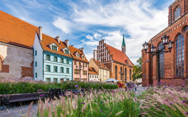 Historic European street scene with colorful gabled houses, a brick church with a green spire, and vintage lampposts under a vibrant blue sky.