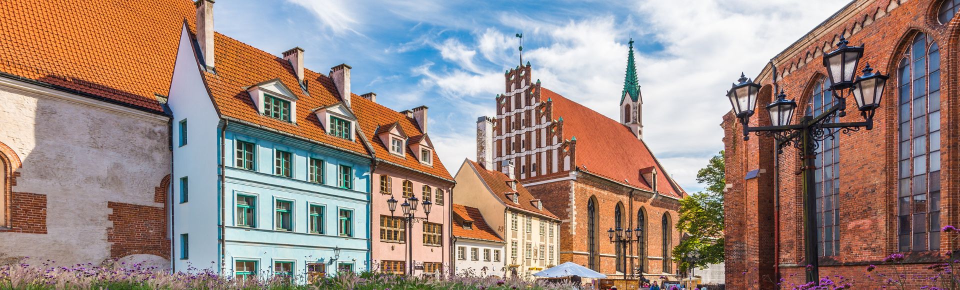 Historic European street scene with colorful gabled houses, a brick church with a green spire, and vintage lampposts under a vibrant blue sky.
