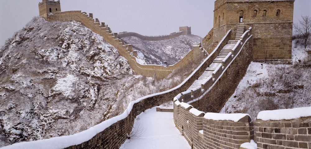 The Great Wall of China in winter, covered in snow. A fortified tower stands on a rugged, snowy mountain ridge under an overcast sky, evoking solitude.