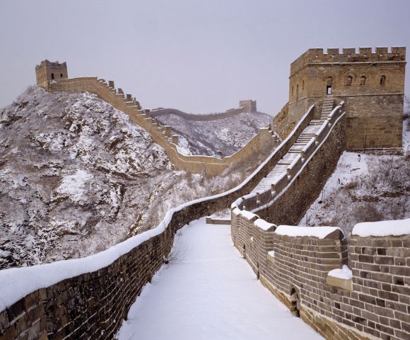 The Great Wall of China in winter, covered in snow. A fortified tower stands on a rugged, snowy mountain ridge under an overcast sky, evoking solitude.