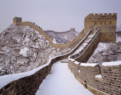 The Great Wall of China in winter, covered in snow. A fortified tower stands on a rugged, snowy mountain ridge under an overcast sky, evoking solitude.