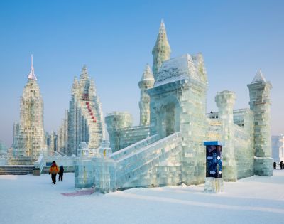 A majestic ice castle with intricate spires stands under a clear blue sky. Two people in winter clothing walk nearby, creating a serene winter scene.