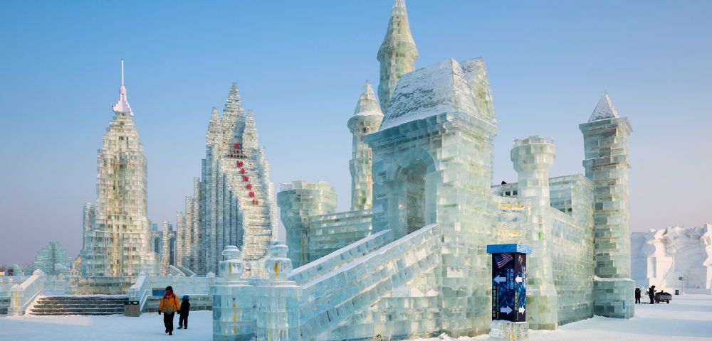 A majestic ice castle with intricate spires stands under a clear blue sky. Two people in winter clothing walk nearby, creating a serene winter scene.