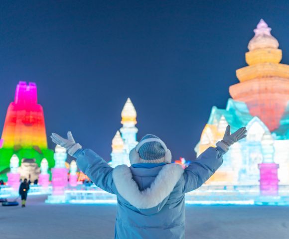 A person in a winter coat stands with arms outstretched, facing illuminated ice sculptures in vibrant colors against a clear night sky, conveying wonder.