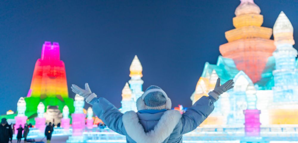 A person in a winter coat stands with arms outstretched, facing illuminated ice sculptures in vibrant colors against a clear night sky, conveying wonder.