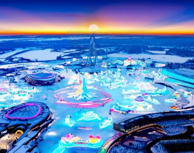 Aerial view of an illuminated winter festival at sunset. Vibrant neon lights form intricate designs on snow, surrounded by dark, snowy fields. Energetic and magical.