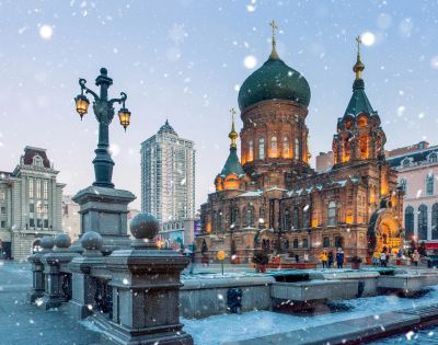 Snow falls gently on a beautifully lit, historic, onion-domed church surrounded by ornate street lamps. The scene evokes a serene winter evening.