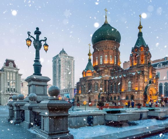 Snow falls gently on a beautifully lit, historic, onion-domed church surrounded by ornate street lamps. The scene evokes a serene winter evening.
