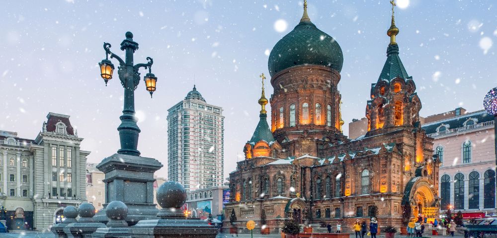 Snow falls gently on a beautifully lit, historic, onion-domed church surrounded by ornate street lamps. The scene evokes a serene winter evening.