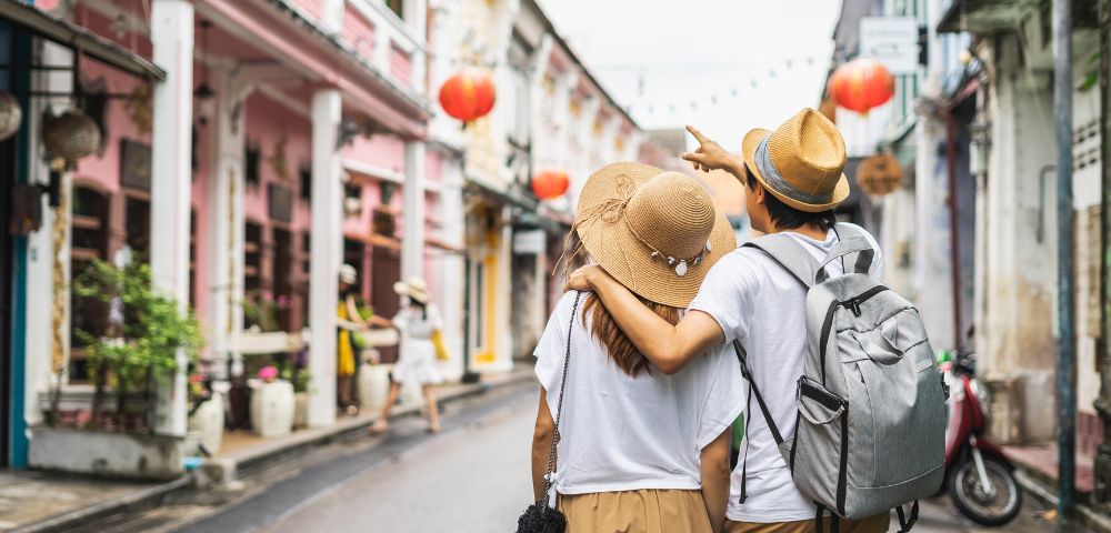A couple wearing summer hats stroll down a colorful street lined with shops. Red lanterns hang above, creating a vibrant, cheerful atmosphere.