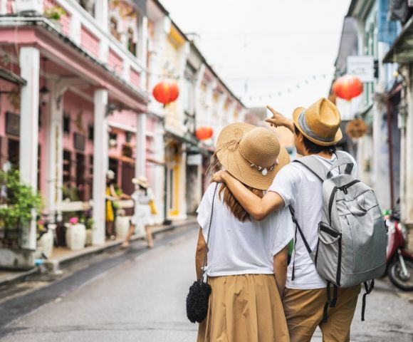 A couple wearing summer hats stroll down a colorful street lined with shops. Red lanterns hang above, creating a vibrant, cheerful atmosphere.