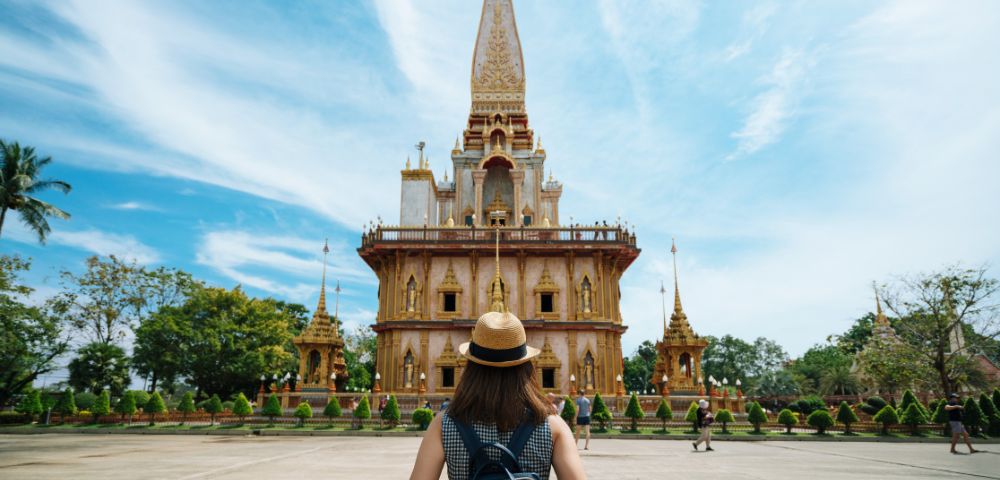 A person with a backpack stands before an ornate, golden temple against a bright blue sky, conveying a sense of awe and exploration.