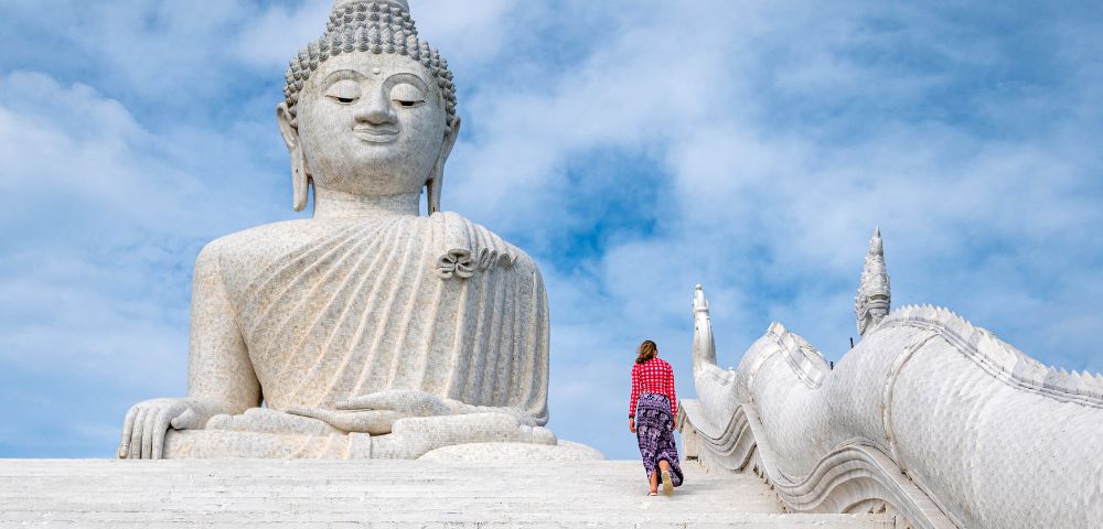 A woman in colorful attire walks up white steps towards a massive stone Buddha statue under a bright blue sky, conveying serenity and reverence.