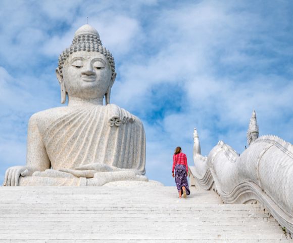 A woman in colorful attire walks up white steps towards a massive stone Buddha statue under a bright blue sky, conveying serenity and reverence.