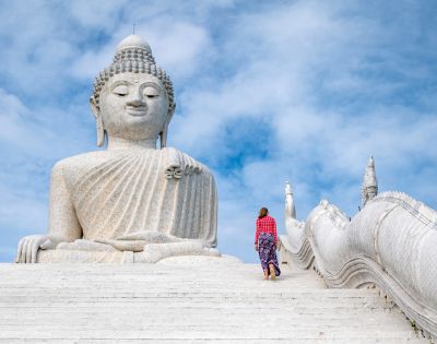 A woman in colorful attire walks up white steps towards a massive stone Buddha statue under a bright blue sky, conveying serenity and reverence.