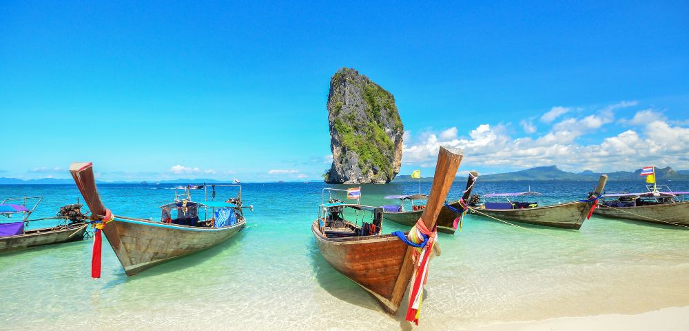 Five colorful long-tail boats are anchored on a sunny beach with clear turquoise water, a tall rock island in the distance, and a vibrant blue sky above.