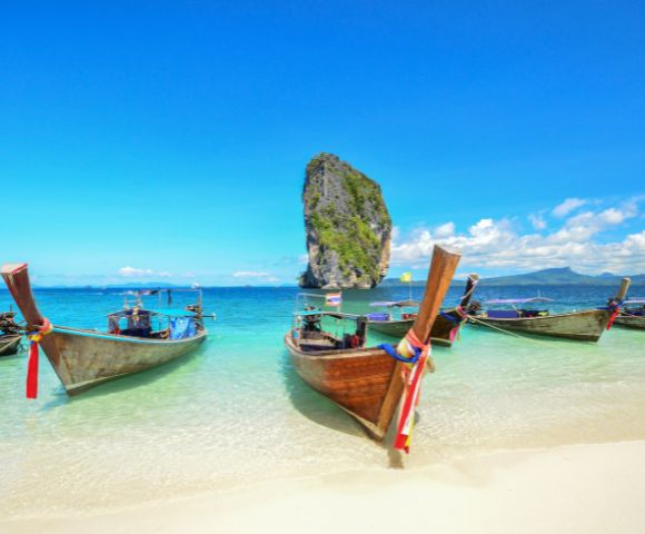 Five colorful long-tail boats are anchored on a sunny beach with clear turquoise water, a tall rock island in the distance, and a vibrant blue sky above.