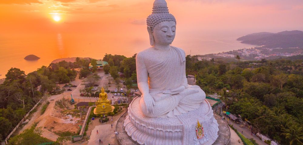 Aerial view of a large white Buddha statue at sunset, sitting serenely on a hill with trees and ocean in the background, exuding tranquility.