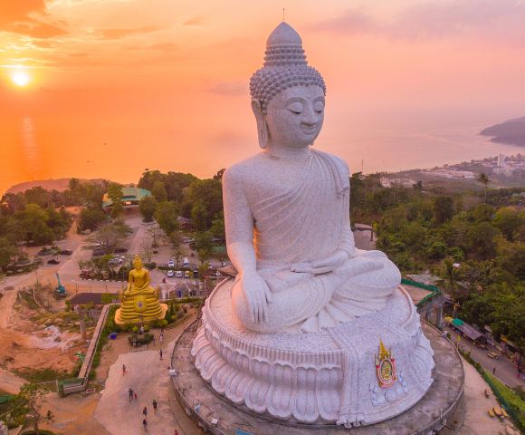 Aerial view of a large white Buddha statue at sunset, sitting serenely on a hill with trees and ocean in the background, exuding tranquility.