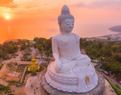 Aerial view of a large white Buddha statue at sunset, sitting serenely on a hill with trees and ocean in the background, exuding tranquility.
