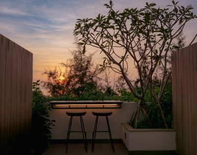 Rooftop patio at sunset with two bar stools facing a wooden counter. Framed by trees and warm light, the scene conveys tranquility and relaxation.