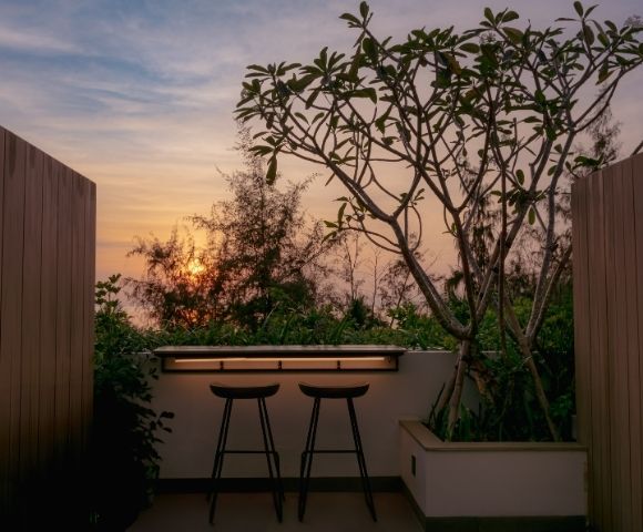 Rooftop patio at sunset with two bar stools facing a wooden counter. Framed by trees and warm light, the scene conveys tranquility and relaxation.