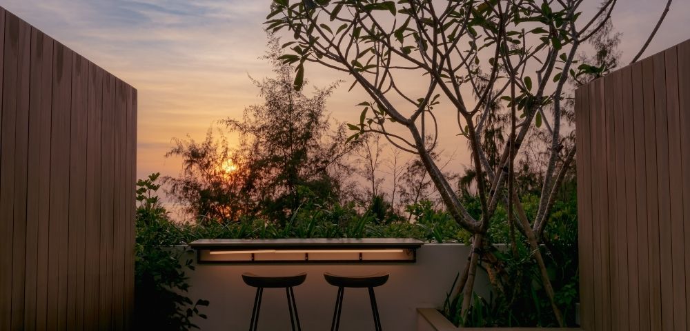 Rooftop patio at sunset with two bar stools facing a wooden counter. Framed by trees and warm light, the scene conveys tranquility and relaxation.