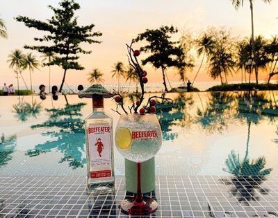Bottle of Beefeater gin and cocktail glass on poolside table at sunset. Palm trees reflect in the water, creating a tranquil, tropical vibe.