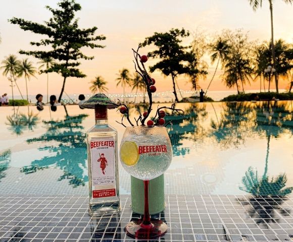 Bottle of Beefeater gin and cocktail glass on poolside table at sunset. Palm trees reflect in the water, creating a tranquil, tropical vibe.