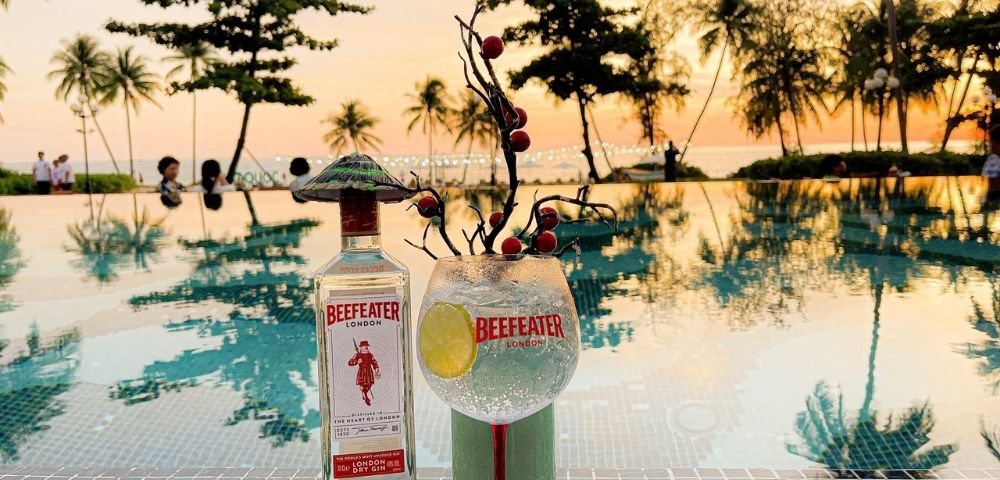 Bottle of Beefeater gin and cocktail glass on poolside table at sunset. Palm trees reflect in the water, creating a tranquil, tropical vibe.