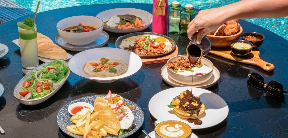 A vibrant brunch spread by a pool includes salads, pasta, pancakes, pastries, and colorful drinks. A hand pours syrup over pancakes, creating a relaxed, indulgent vibe.